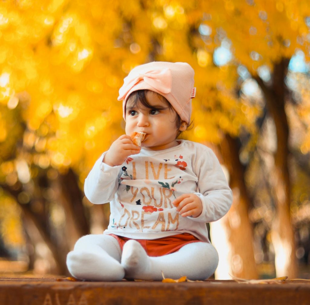 A cute baby girl sitting on a bench enjoying a sunny fall day in a park.