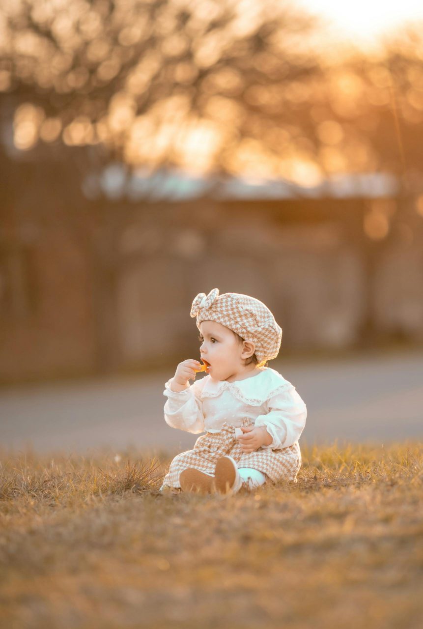 Adorable baby girl in checkered outfit sitting on grass in autumn sunset.